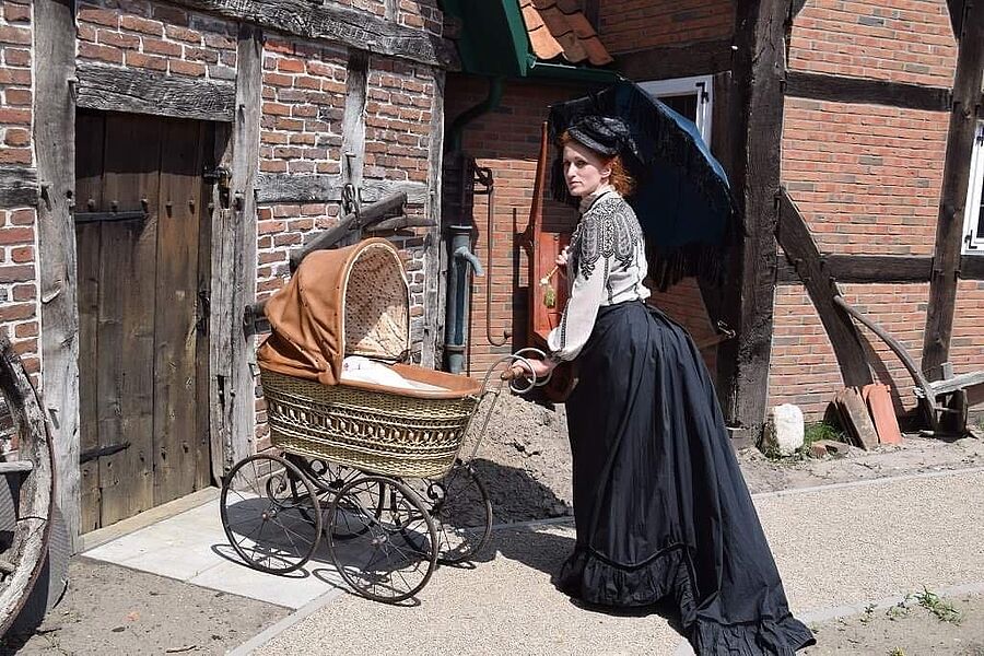 Frau in historischem Kleid schiebt einen Kinderwagen ins Gebäude