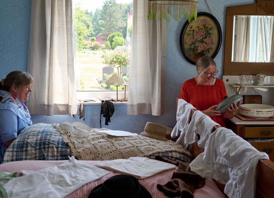 Blick in ein Schlafzimmer, eine Person sitzt im Bett, eine andere auf einem Stuhl davor 
