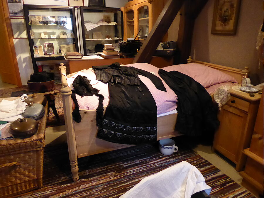 Blick in ein Wohnzimmer mit Bett, auf dem Kleidungsstücke liegen